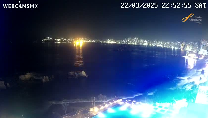 A nighttime view of a city illuminated across a dark body of water, with a beach and a pool visible in the foreground.