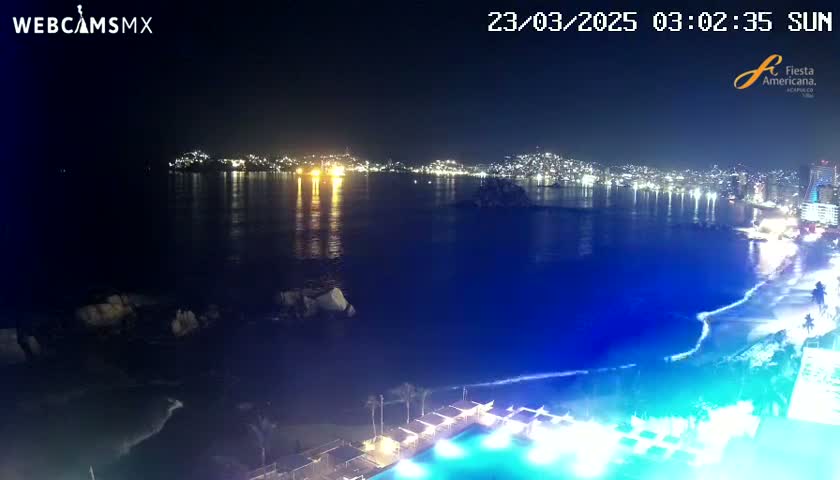 A nighttime coastal scene shows a city's lights across a dark blue ocean, with a beach and illuminated pool in the foreground.