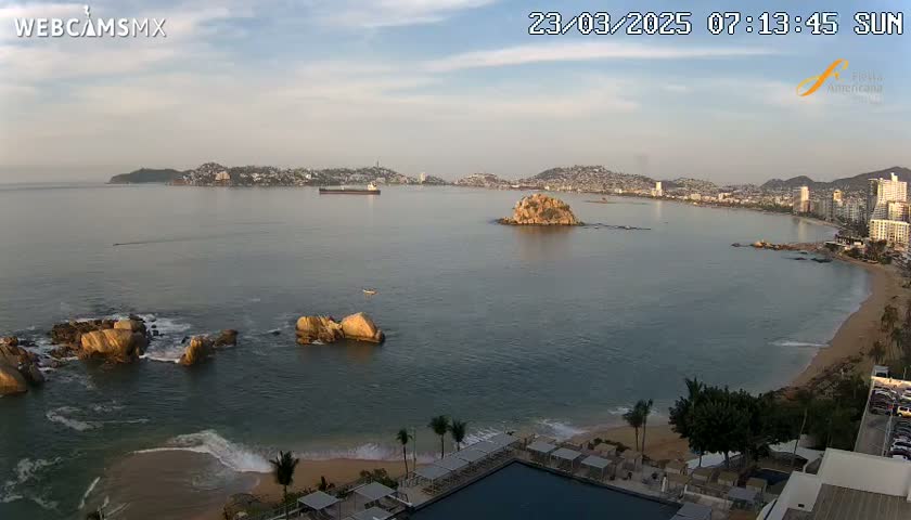 A calm, sunny morning view of a bay with a city in the distance, rocky outcrops in the foreground, and a sandy beach with a resort partially visible.