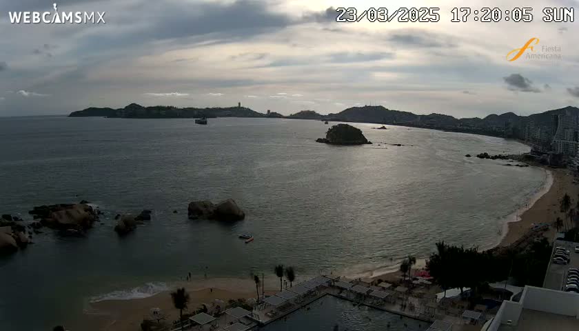 A mostly cloudy sunset over a bay with a beach, rocky outcroppings, and distant buildings.