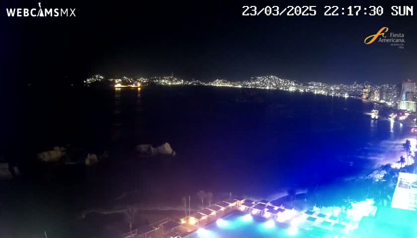 A nighttime view of a city skyline across a dark body of water, with a brightly lit pool and beach area in the foreground.