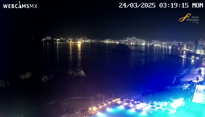 A nighttime view of a city's illuminated coastline across a calm, dark ocean, with a resort pool and beach visible in the foreground.