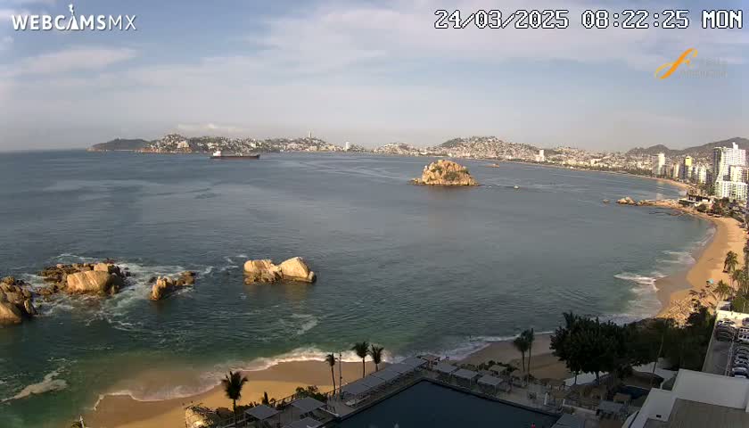A mostly sunny day reveals a wide expanse of ocean, rocky outcrops, a sandy beach, and a city skyline in the distance.