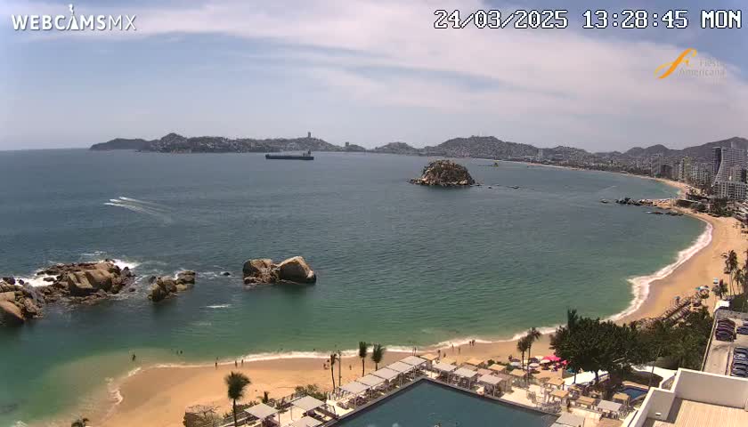 An expansive, sandy beach curves around a calm, teal ocean under a mostly sunny sky, with rocky outcrops and a distant cityscape visible.