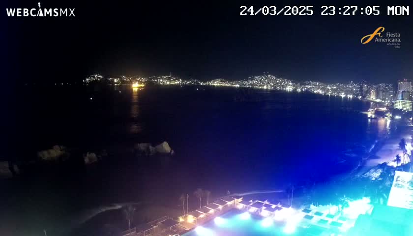 A nighttime coastal view shows a city's lights across a dark bay, with a brightly lit pool and beach area in the foreground.