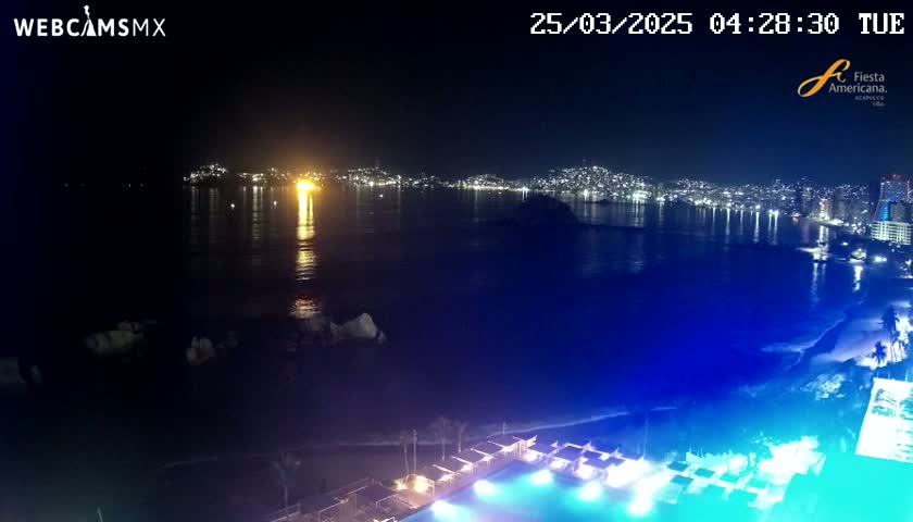 A nighttime view of a city's lights across a dark body of water, with a brightly lit pool and beach in the foreground.