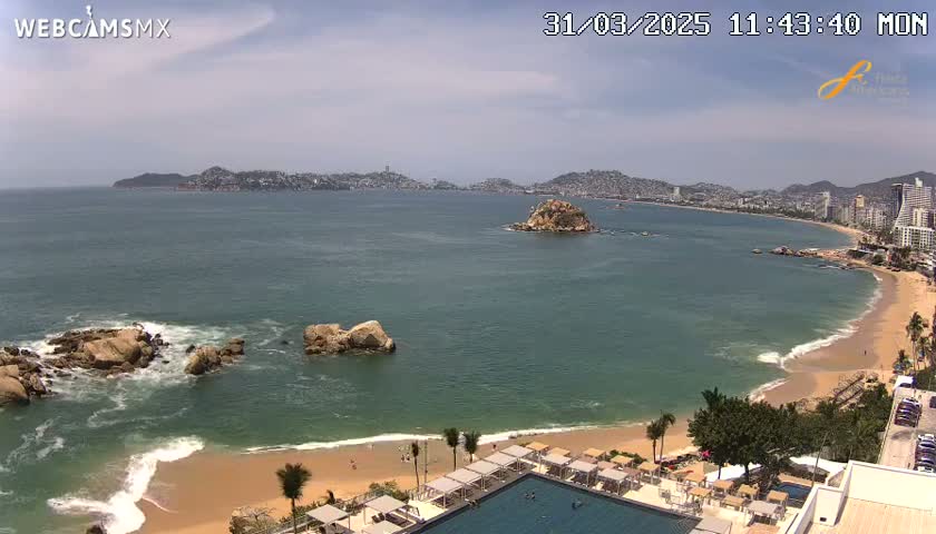 A beach with calm waves and a resort with a pool is situated near a rocky coastline overlooking a bay with a distant city under a mostly clear sky.