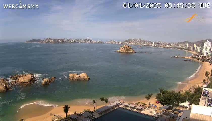 A sunny coastal scene shows a city skyline across a calm bay with a sandy beach in the foreground, featuring a resort with a pool.