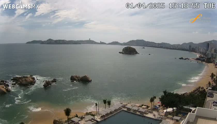 A partly cloudy day reveals a sweeping ocean vista, featuring a sandy beach, rocky outcrops, distant hills, and a resort with a swimming pool.