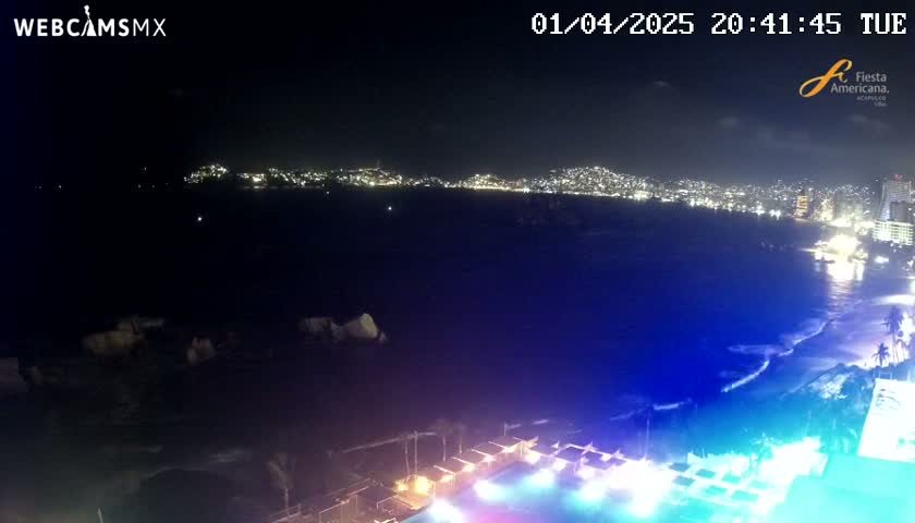 A nighttime coastal view shows a city's lights across a dark body of water, with a beach and illuminated structures in the foreground under a clear sky.
