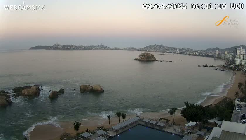 A calm, pale-pink dawn illuminates a coastal scene with a sandy beach, a resort pool, rocky outcroppings in the ocean, and a city skyline in the distance.