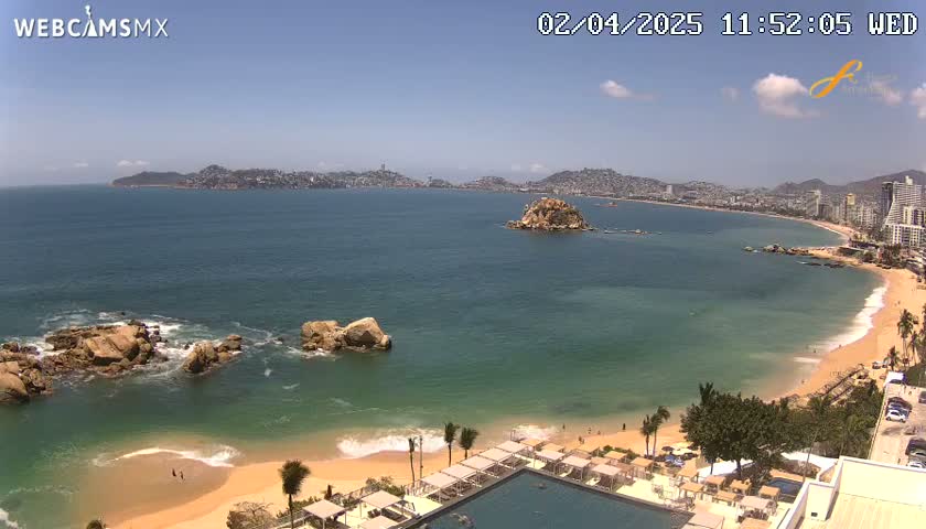 A sunny, panoramic view shows a calm ocean bay with a sandy beach, rocky outcrops, and a distant city under a mostly clear sky.