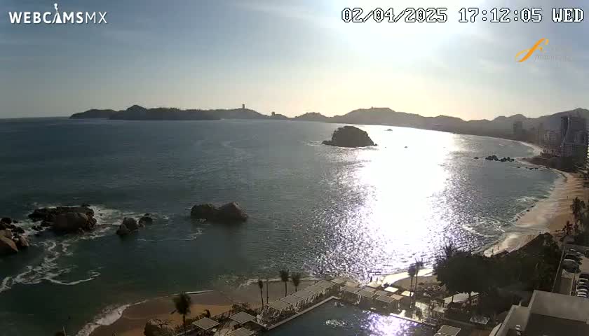 A sunny coastal scene shows a calm ocean reflecting sunlight, rocky outcrops near a sandy beach, and distant hills under a clear sky.