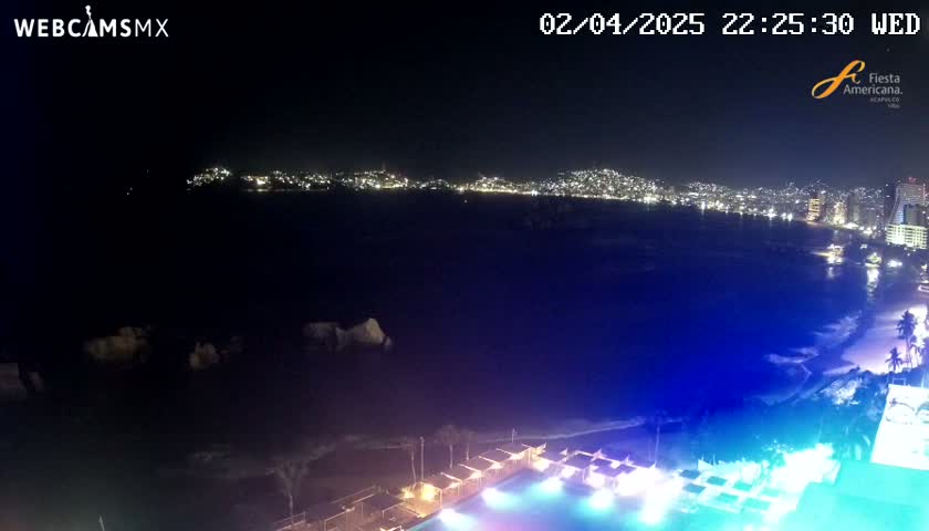 A nighttime, coastal view shows a city skyline across a dark body of water, with a brightly lit pool and beachside cabanas in the foreground.