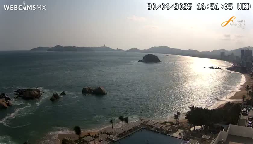 A sunny day reveals a wide ocean bay with rocky islands, a sandy beach, and a resort with a swimming pool.