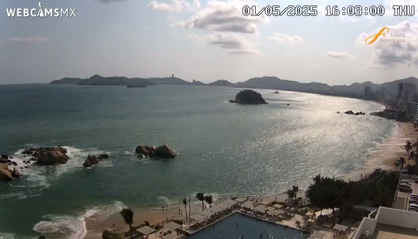 A partly sunny day reveals a tranquil ocean scene with rocky outcroppings near a beach and resort, featuring a swimming pool.