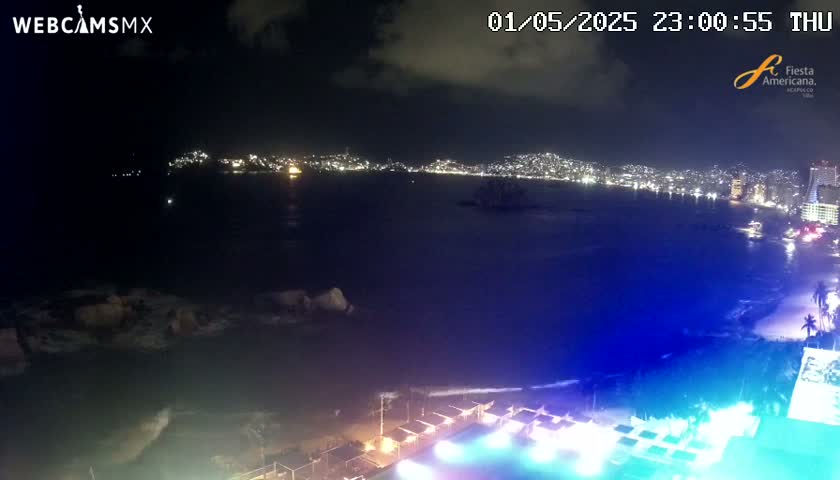 A nighttime coastal scene shows a city brightly lit across a dark body of water with gently lapping waves on a beach in the foreground.