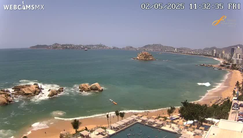 A sunny beach with calm, clear water and rocky outcrops, overlooks a bay with a distant city skyline.