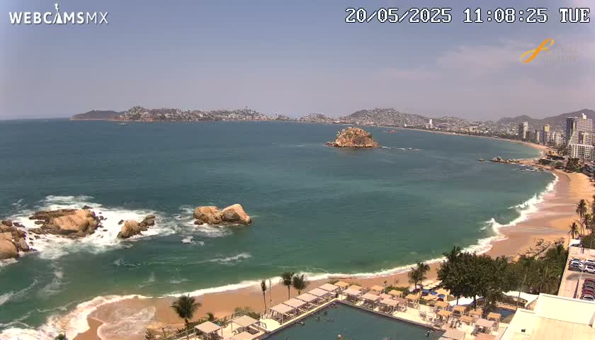 A sunny day shows a sweeping view of a coastline with turquoise water lapping a sandy beach, rocky outcroppings, and a resort with a pool in the foreground, backed by a city on a hill.