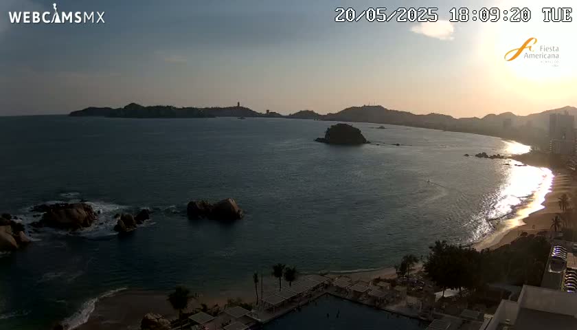 A sunset over a calm ocean, illuminating a sandy beach with a resort in the foreground and rocky islands in the bay.