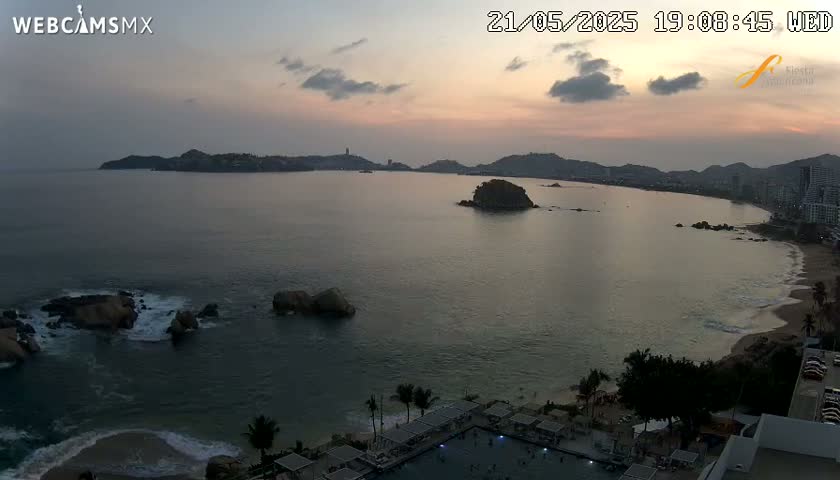 An ocean bay at sunset, with a beach, rocky outcrops, and a coastal town visible in the distance under a partly cloudy sky.