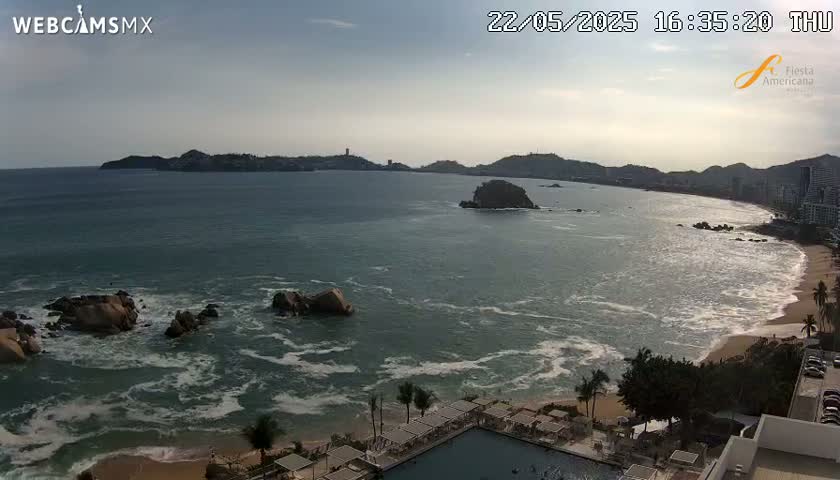 An ocean scene with moderate waves shows a beach with a resort and several rocky outcrops, under a partly cloudy sky.
