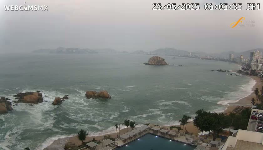 A hazy, overcast view of a rocky coastline with a beach, ocean waves, and a resort pool in the foreground.