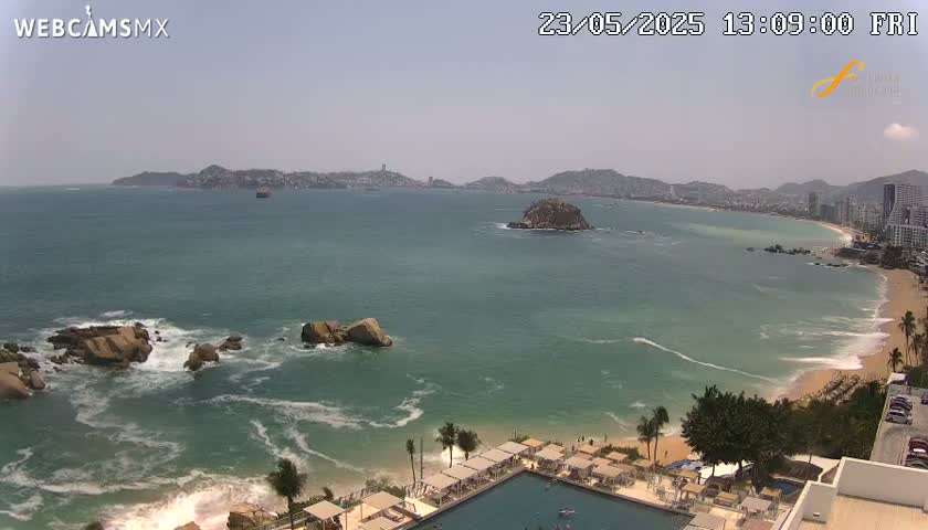 A sunny, high-angle view shows a coastal scene with a calm ocean, sandy beach, rocky outcroppings, and a resort with a swimming pool.