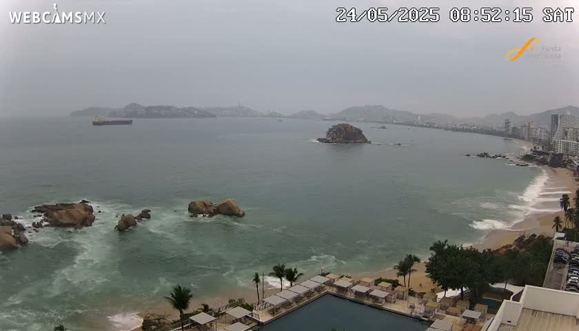 A hazy coastal scene shows a calm bay with a cargo ship, rocky outcroppings, a sandy beach with waves, and a resort with a swimming pool.