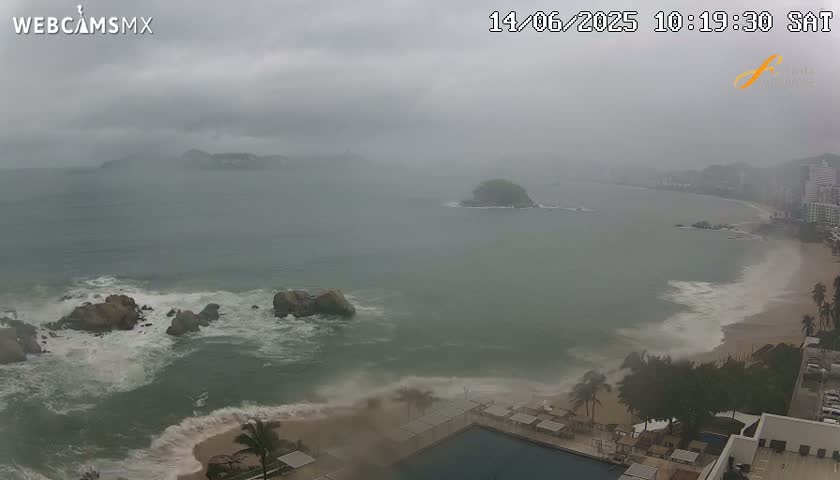 A coastal scene shows a hazy, overcast day with waves crashing on a sandy beach near rocky outcrops and a small island, all under a heavy grey sky.