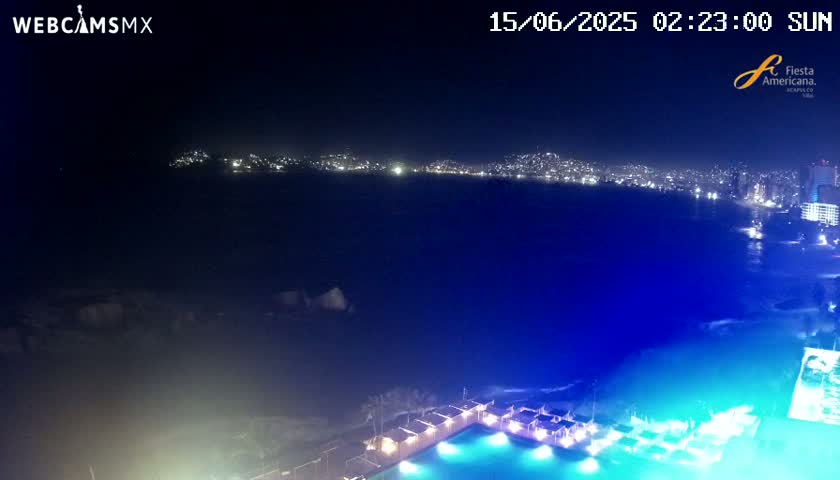 A nighttime, seaside view shows a city skyline across a dark body of water, with a brightly lit pool and surrounding structures in the foreground.