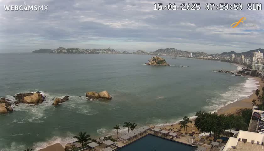 A coastal scene shows a city on a hillside overlooking a bay with a small island, rocky shoreline, and a sandy beach under a mostly cloudy sky.