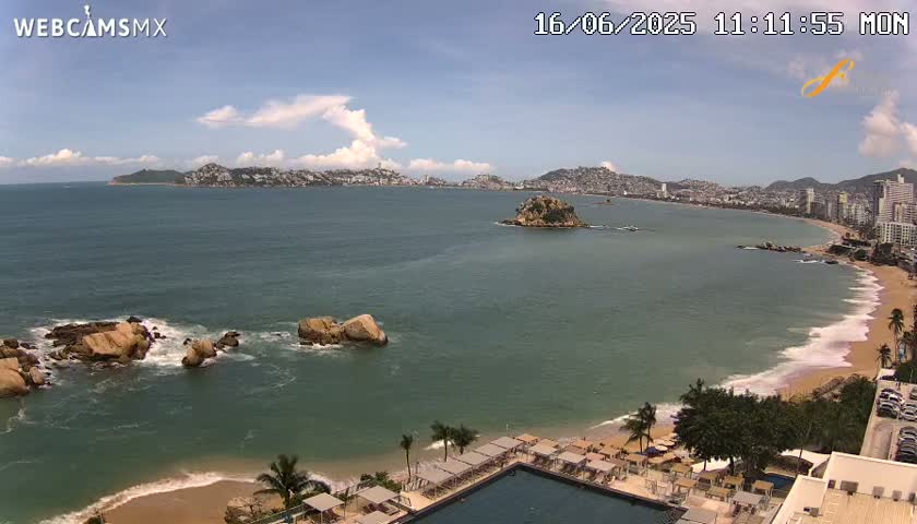 A partly cloudy day shows a coastal scene with a sandy beach, calm ocean waters, rocky outcrops, and a distant city skyline.