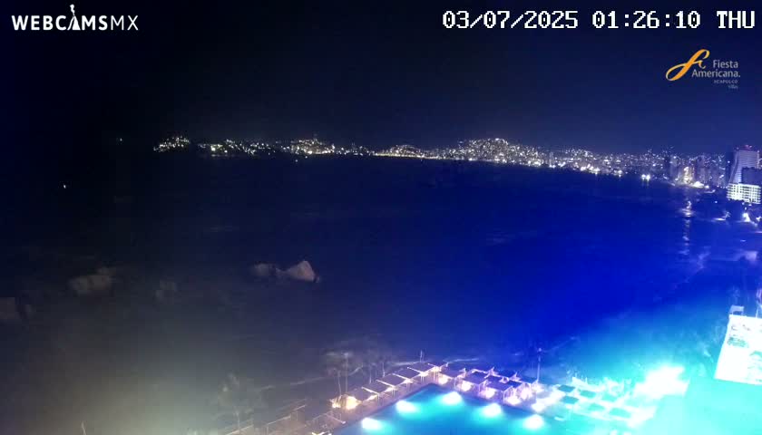 A nighttime view of a city skyline across a dark blue ocean, with a brightly lit pool and cabanas in the foreground.