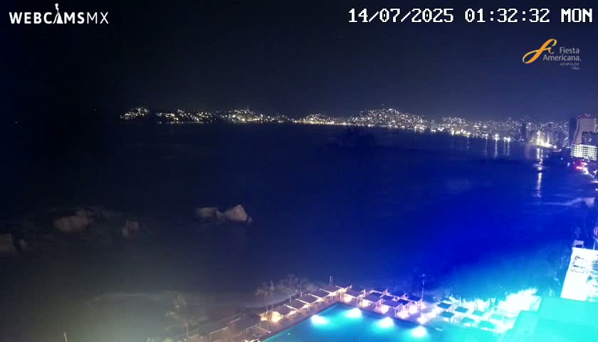 A nighttime, seaside view shows a city skyline across a dark body of water, with a brightly lit pool and surrounding structures in the foreground.