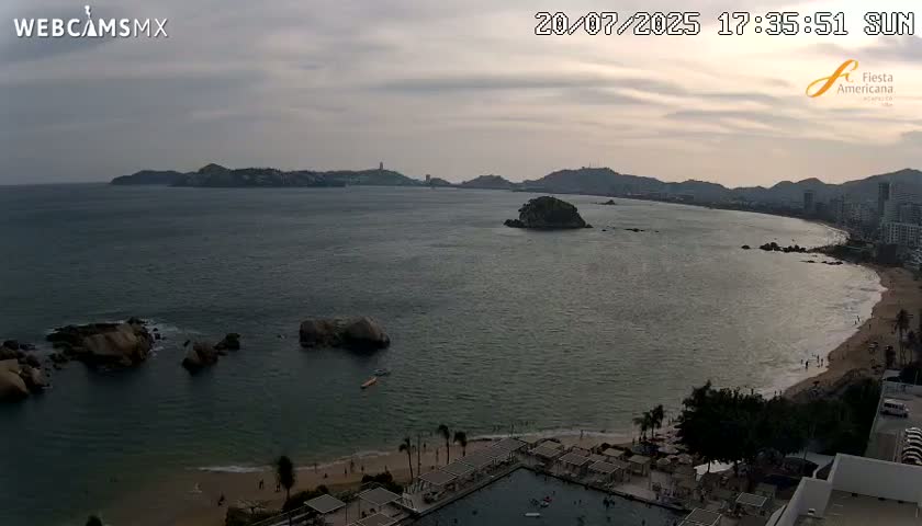 An overcast sunset view of a calm ocean bay with a beach, rocky outcroppings, and distant hills.