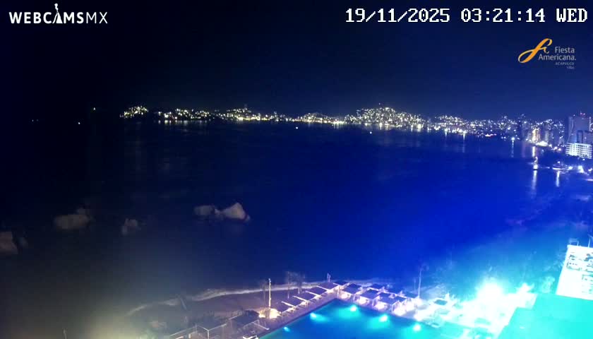 On a clear night, an aerial view captures a brightly lit city sprawling along a curving coastline across a dark bay, with an illuminated beach, swimming pool, and cabanas visible in the foreground, and rocks in the water.