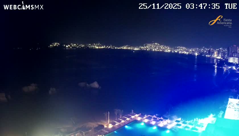 A nighttime aerial view overlooks a wide bay with a brightly illuminated city skyline stretching along the distant shore, while a resort's glowing swimming pool and covered cabanas line the immediate foreground under clear, dark skies.