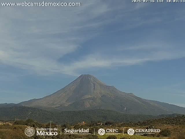 A large, dark-grey volcano is seen on a mostly clear day with a few wispy clouds.