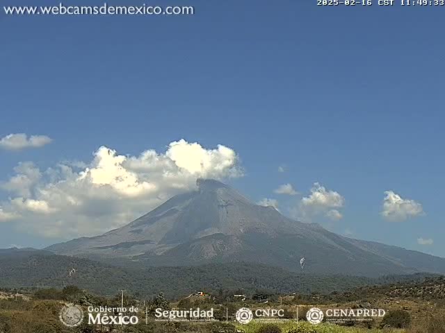 A large, dark-grey volcano is visible under a mostly clear blue sky with scattered cumulus clouds.