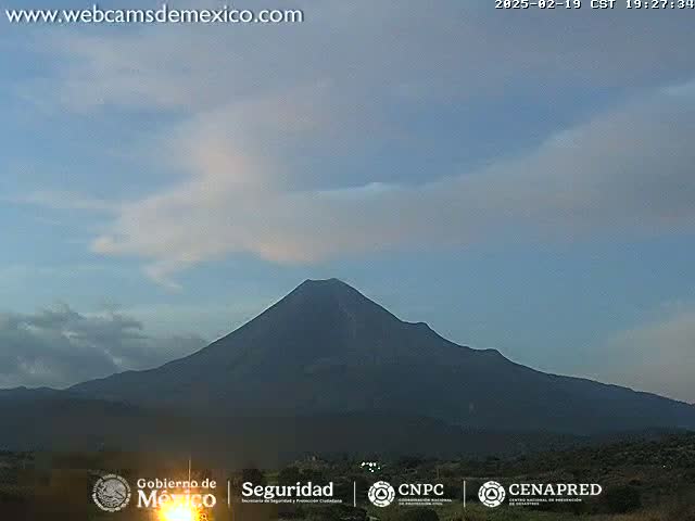 A large, dark volcano is silhouetted against a pale blue sky with thin clouds at dusk.
