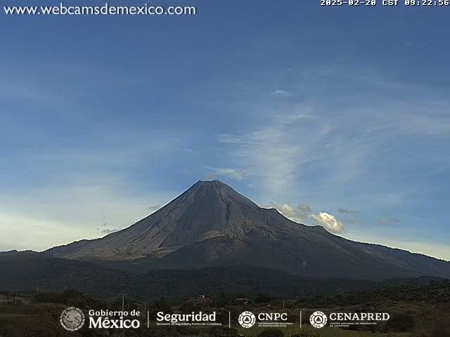 A large, dark volcano is visible under a mostly clear, light blue sky with a few wispy clouds.