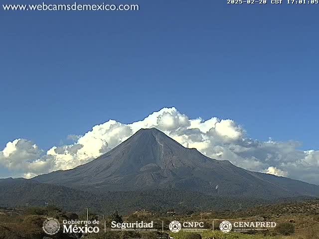 A large, dark-colored volcano is visible under a mostly clear blue sky with puffy white clouds partially obscuring the peak.