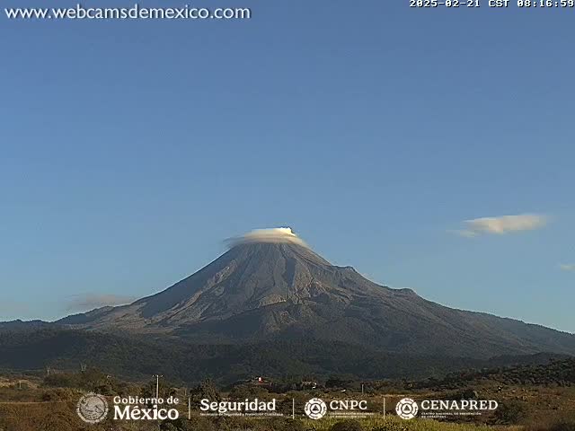 A large, dark-colored volcano with a cap of clouds is visible under a clear blue sky.