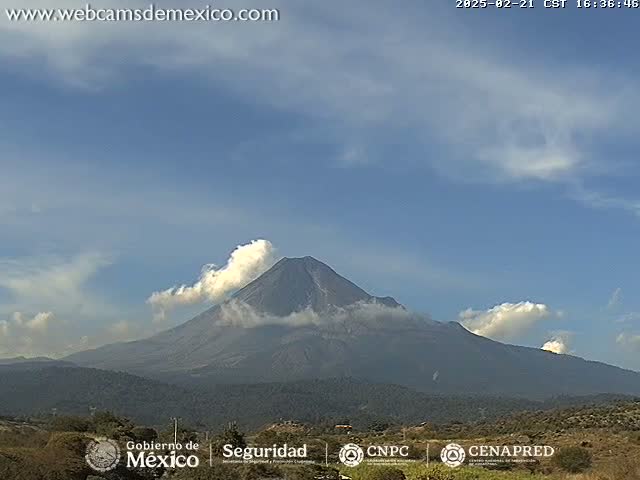 A large, dark volcano with a plume of white smoke is visible under a mostly clear, light blue sky.