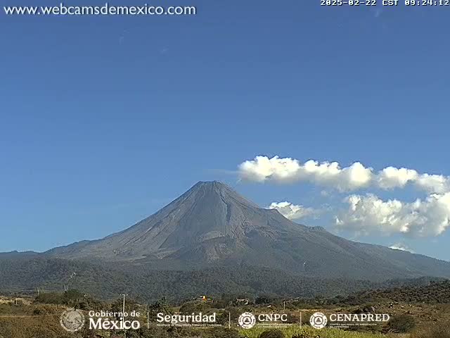 A large, dark-colored volcano is seen on a mostly clear day with a few wispy clouds near its peak.