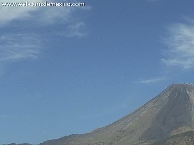 A large, light gray volcano is visible under a mostly clear, light blue sky with a few scattered clouds.