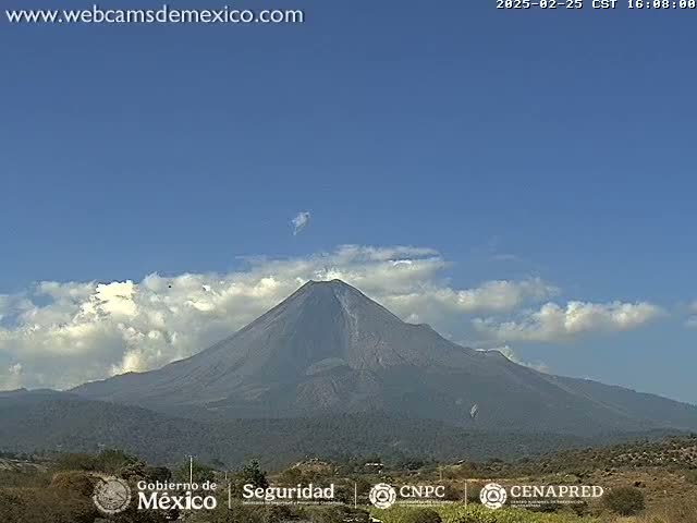 A large, dark volcano is visible under a mostly clear blue sky with some fluffy white clouds.