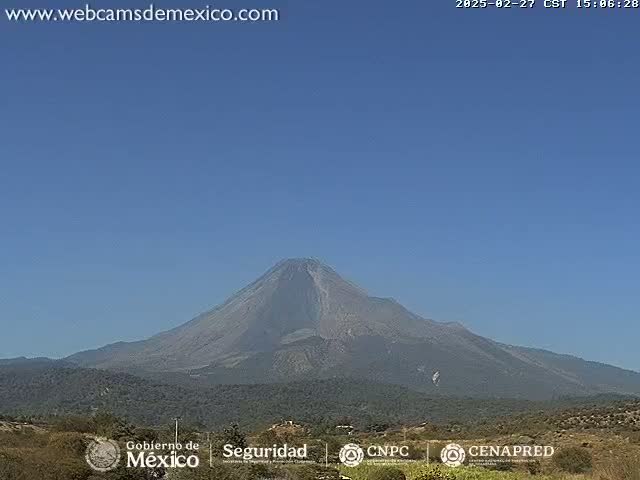 A large, dark-grey volcano is visible on a clear, sunny day, set against a light-blue sky, with low-lying vegetation in the foreground.