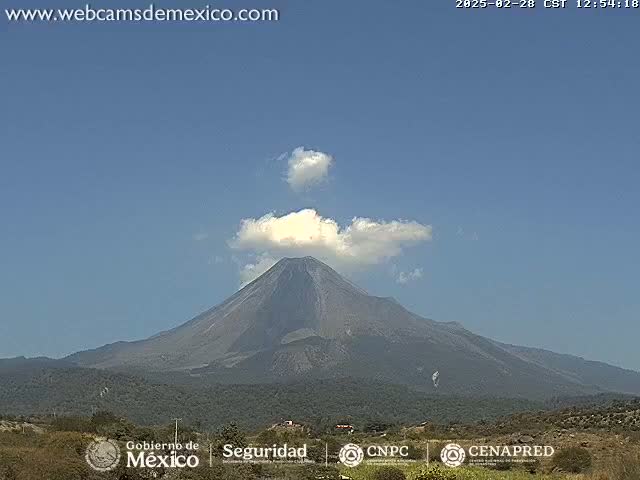A large, dark-colored volcano with a small plume of white smoke erupting from its peak is visible under a clear blue sky.